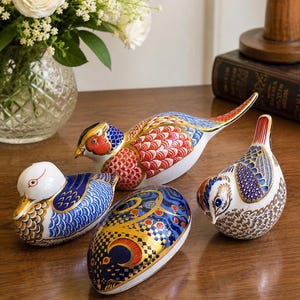 Könnte beinhalten: Eine Sammlung von vier Porzellanfiguren, bestehend aus drei Vögeln und einem eiförmigen Objekt. Die Stücke sind mit komplizierten Mustern in Blau, Rot, Gold und Weiß verziert und zeigen detaillierte Federmotive sowie geometrische Designs.