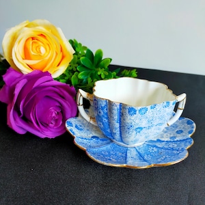 May include: A blue and white teacup and saucer with a floral pattern and gold trim. The cup has two handles and a scalloped edge. The saucer is also scalloped and has a floral pattern.