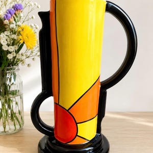 Peut inclure: Un grand pichet en céramique arborant un motif de soleil rayonnant aux tons jaune, orange et rouge. L'objet présente une base noire brillante, une anse noire assortie et des contours noirs soulignant les rayons. Il repose sur un socle circulaire.