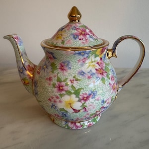 May include: A porcelain teapot featuring a vibrant floral pattern with pink, white, and blue flowers over a light green crackle glaze background. The teapot includes a matching lid with a gold-coloured finial, a curved spout, and a gold-trimmed handle.