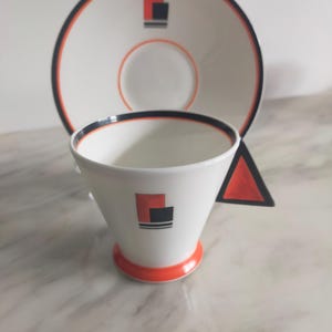 Op de afbeelding: Een witte keramische theekop en schotelset met een geometrisch ontwerp in zwart, oranje en rood. De kop heeft een brede, uitlopende vorm en een klein handvat. De schotel heeft een zwarte rand en een eenvoudig geometrisch ontwerp in het midden.