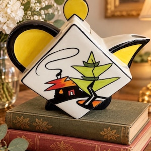 Puede incluir: Una tetera de cerámica en forma de diamante con base blanca y un gráfico pintado a mano que muestra una casa y un árbol estilizado. La tetera tiene un asa negra, un remate de tapa circular amarillo y detalles amarillos en el pico y el asa.