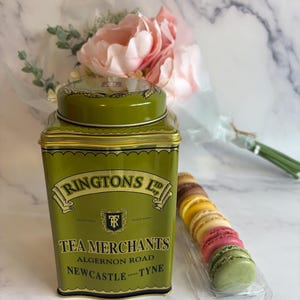 Peut inclure: Une boîte à thé Ringtons Ltd. vert olive avec des bordures dorées et du texte, à côté d'un récipient en plastique transparent contenant des macarons colorés et un bouquet de fleurs roses.