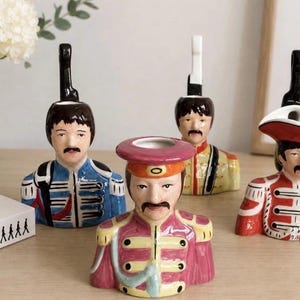 May include: Four ceramic busts depicting members of The Beatles in colorful military-style uniforms. Each figure features a mustache, dark hair, and a hollow top opening. The uniforms are blue, pink, yellow, and red, with intricate detailing.