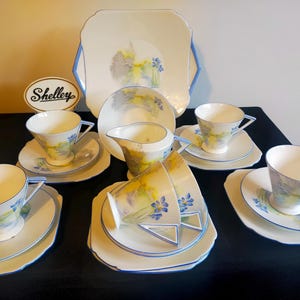 May include: A vintage tea set with a floral design. The set includes a teapot, creamer, sugar bowl, and six teacups and saucers. The teacups and saucers have a scalloped edge and are decorated with a delicate floral pattern in shades of blue, yellow, and grey. The teapot, creamer, and sugar bowl are also decorated with the same floral pattern. The set is made by Shelley.