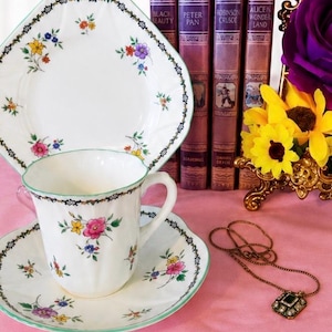 May include: A white porcelain teacup and saucer set featuring a delicate floral pattern with pink, yellow, and blue flowers, accented by a black and green border. A matching plate stands behind the cup, alongside four purple books and a necklace.