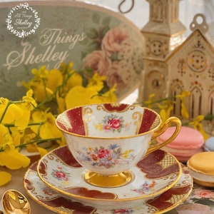 May include: A white porcelain teacup and saucer set featuring a deep red border with gold filigree accents and a central floral bouquet of pink, blue, and yellow flowers. The teacup has a gold-trimmed handle and a gold-finished pedestal base.