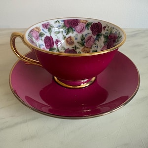 May include: A magenta tea cup and saucer set featuring a gold-trimmed handle, rim, and base. The interior of the cup is decorated with a colourful floral pattern of pink, red, and yellow roses against a white background, contrasting with the solid magenta exterior.