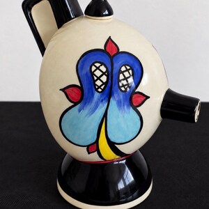 Könnte beinhalten: Eine Keramik-Teekanne mit einem cremefarbenen, kugelförmigen Körper, einem schwarzen Griff, einem schwarzen Ausguss und einem schwarzen runden Sockel. Der Körper zeigt ein handgemaltes Design mit blauen, gelben und roten Akzenten.