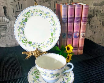 Könnte beinhalten: Ein blaues und weißes Tee-Set mit Untertasse und einem Blumenmuster. Das Set steht auf einer schwarzen Oberfläche mit einem goldenen Ständer und einem Stapel Bücher dahinter.