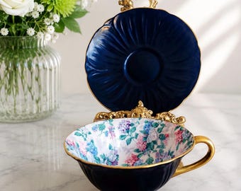 May include: A dark navy blue teacup and saucer set featuring a gold rim and handle. The interior of the teacup is decorated with a colorful floral pattern of blue, pink, and purple hydrangea blossoms. The saucer is displayed vertically behind the cup.