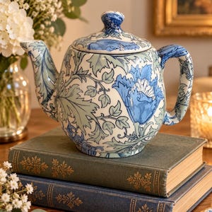 May include: A ceramic teapot featuring a white base with a repeating blue and sage green floral and leaf pattern. The teapot has a curved spout, a sturdy handle, and a matching lid with a small knob handle on top, all decorated in the same motif.