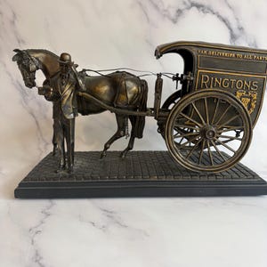 Peut inclure: Sculpture en bronze d'une charrette tirée par un cheval avec un conducteur. La charrette porte l'inscription "RINGTONS" et le texte "VAN DELIVERIES TO ALL PARTS". La sculpture est posée sur une base rectangulaire noire.