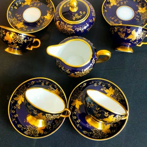 Puede incluir: Juego de té completo de porcelana azul marino que incluye tazas, platillos, azucarero y jarra de crema. El juego está adornado con patrones de hojas doradas y detalles dorados. Las tazas y platillos tienen un interior blanco.