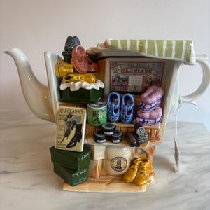 以下が含まれることがあります： 靴屋の形をしたセラミックティーポット。ミニチュアの靴、ブーツ、アクセサリーが特徴です。ティーポットは白で、靴屋のファサードがあり、「The Hygienic Boots & Shoes C.J. Clark」と書かれた看板が付いています。