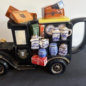 May include: A black ceramic teapot shaped like a vintage delivery van, featuring gold-colored headlights and wheels. The side displays shelves holding miniature blue and white porcelain tea items, green boxes, and red Ringtons Tea packaging.