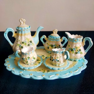 Peut inclure: Un service à thé miniature comprenant une théière, un sucrier, une crémière et deux tasses sur soucoupes, disposés sur un plateau festonné. L'ensemble est peint en bleu clair, crème et jaune, avec des motifs floraux. La théière a un couvercle en forme de couronne.