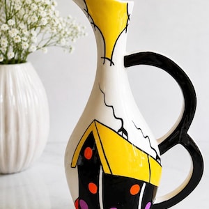 Peut inclure: Un grand pichet en céramique à base blanche, doté d'une anse noire et d'un bec verseur jaune. Le corps est orné d'une illustration graphique représentant une maison avec un toit jaune, des murs noirs, des accents circulaires orange et une base rouge.