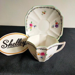 May include: A white teacup and saucer with a green trim and floral pattern. The saucer is square with rounded corners and the teacup has a scalloped edge. The teacup and saucer are made by Shelley.