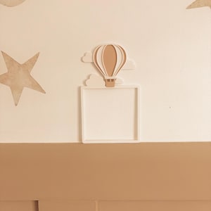 May include: A white square frame with a beige hot air balloon and cloud design. The wall decor also features beige stars and clouds. The color palette is neutral, with beige and white tones, creating a soft, whimsical aesthetic.