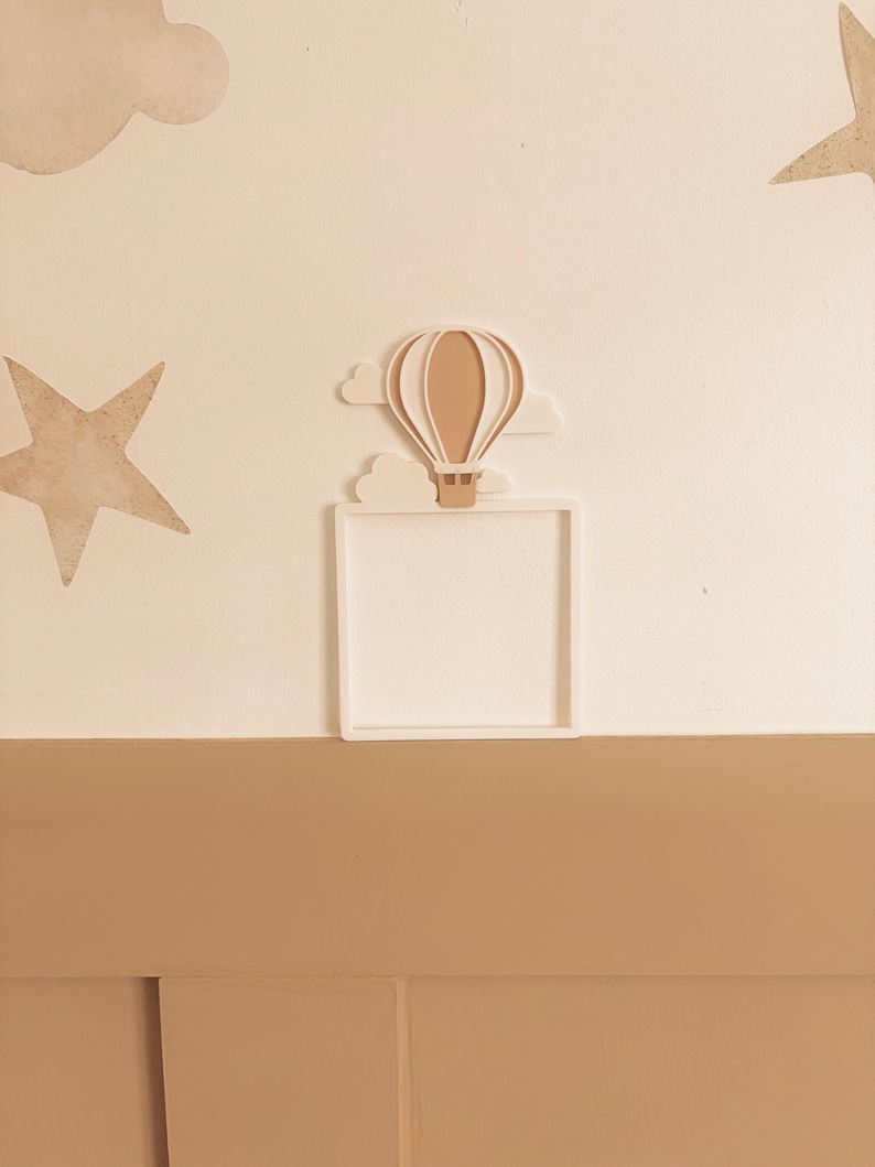 May include: A white square frame with a beige hot air balloon and cloud design. The wall decor also features beige stars and clouds. The color palette is neutral, with beige and white tones, creating a soft, whimsical aesthetic.