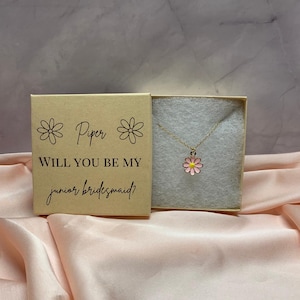 Puede incluir: Un collar dorado con un colgante de margarita rosa se presenta en una caja de regalo. La caja beige tiene el texto "Piper, WILL YOU BE MY junior bridesmaid?" y gráficos de margaritas. El collar está sobre tela rosa.