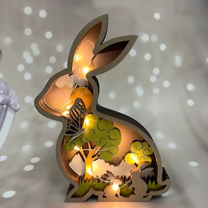 May include: A wooden rabbit cutout with a layered design, featuring a butterfly, a tree, and a smaller rabbit. The cutout is illuminated with warm white LED lights, creating a whimsical and festive atmosphere.