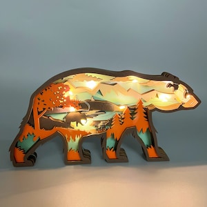 Puede incluir: Una silueta de oso de madera con un diseño en capas, que presenta una escena de bosque con árboles, montañas y un río. El oso está iluminado con luces LED blancas cálidas.