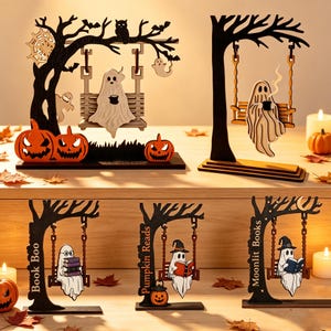 Pode incluir: Uma coleção de cinco decorações de mesa em madeira para o Halloween com fantasmas em baloiços. Os designs incluem fantasmas com canecas, livros e abóboras. As árvores são pretas e o texto diz "BOOK BOO", "PUMPKIN READS" e "MOONLIT BOOKS".