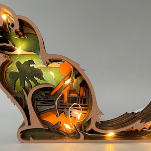 May include: A wooden cat figurine with a layered design, featuring a tropical scene inside. The cat is sitting with its tail curled around its body. The scene includes palm trees, a beach, and a sunset. The figurine is illuminated with warm white LED lights.