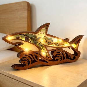 Puede incluir: Escultura de tiburón de madera con luces LED internas. El tiburón es marrón con un diseño detallado, incluyendo un interior en capas con acentos verdes y beige. La escultura se asienta sobre una base en forma de ola, y las luces iluminan la forma del tiburón.