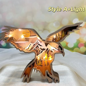 Könnte beinhalten: Eine Holzskulptur eines Adlers mit einem geschichteten Design, beleuchtet von warmweißen LED-Lichtern. Die Flügel des Adlers sind weit ausgebreitet, und das Design zeigt eine Bergszene mit Bäumen und einer Sonne. Die Skulptur ist mit "Style A+Light" beschriftet.