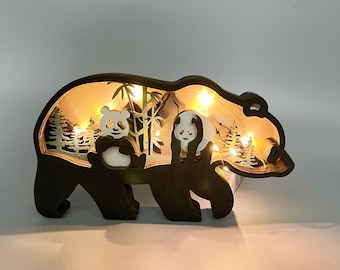 Puede incluir: Una silueta de oso de madera en forma de panda con una función de iluminación. El oso está recortado para revelar una escena de pandas y bambú en el interior. El oso es negro y la escena está iluminada con luces LED blancas cálidas.