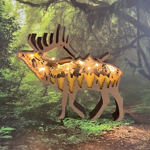 May include: A wooden cutout of an elk with a forest scene inside. The elk is illuminated with warm white fairy lights.