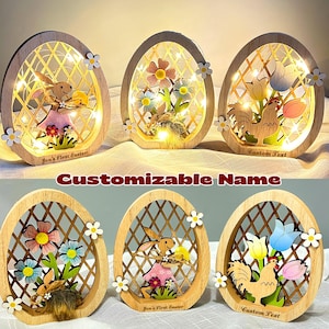 May include: Wooden Easter egg decorations with illuminated interiors. Each egg features a unique design: a bunny, flowers, and a rooster. The eggs are framed with a lattice pattern and adorned with small white flowers. The text "Jon's First Easter" and "Custom Text" are visible.