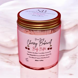 Op de afbeelding: Een roze pot met Cherry Bakewell body butter met een gouden deksel. De pot is gevuld met een roze en wit geklopt product, dat lijkt op slagroom. Het etiket op de pot zegt "Nur Beauty Cherry Bakewell Body Butter" en somt de ingrediënten op. De pot is 250 ml of 8,79 oz.