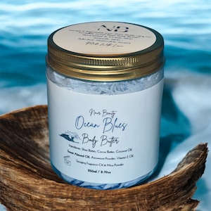 Puede incluir: Un tarro blanco con tapa dorada lleno de manteca corporal azul. La etiqueta dice "Nour Beauty Ocean Blues Body Butter" y enumera los ingredientes: Manteca de karité, Manteca de cacao, Aceite de coco, Aceite de almendras dulces, Polvo de arrurruz, Aceite de vitamina E, Aceite de fragancia y Polvo de mica. El tarro está sobre una superficie de madera.