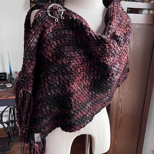 May include: A handmade, fringed shawl in shades of burgundy, black, and gray, displayed on a mannequin. The shawl is secured with a silver-toned, decorative pin. The knitted texture and color palette suggest a cozy, warm accessory, perfect for cooler weather.