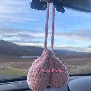 May include: A light pink, plush car accessory shaped like testicles, suspended from a car's rearview mirror. The accessory is made of a soft, textured material. The text "StitchesBySlapp" is visible on the accessory.