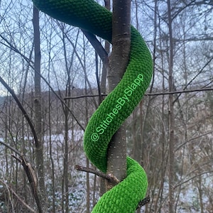Pode incluir: Um brinquedo de cobra de pelúcia verde enrolado em volta de um galho de árvore. A cobra tem uma superfície texturizada e um olho verde. O texto "@StitchesBySlapp" é visível na cobra.