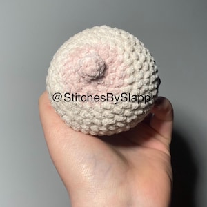 Puede incluir: Una esfera de ganchillo con una superficie texturizada, con un detalle de areola y pezón rosa claro. La esfera se sostiene en una mano. El texto "@StitchesBySlapp" es visible.