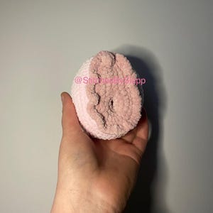 May include: A pink and white crocheted plush toy shaped like a heart. The toy has a small hole in the center. The text "@StitchesBySlapp" is visible on the toy.