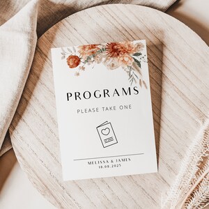 May include: A white sign with the text "Programs" and "Please take one" in black font. The sign is decorated with a floral design in shades of orange, brown, and green. The sign also includes a small icon of a closed book with a heart on the front. The text "Melissa & James" and "18.08.2025" is printed in black at the bottom of the sign.