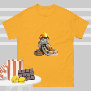 Può includere: Una maglietta gialla con una grafica di un serpente che indossa un casco da cantiere giallo. Il serpente è arrotolato e ha un motivo bianco e nero.