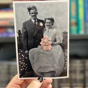 K&ouml;nnte beinhalten: Ein Schwarzwei&szlig;-Hochzeitsfoto eines Paares. Die Braut tr&auml;gt ein wei&szlig;es Kleid und h&auml;lt einen Strau&szlig; rosa Blumen. Der Br&auml;utigam tr&auml;gt einen Anzug und eine Krawatte. Das Foto ist in einem wei&szlig;en Rahmen.