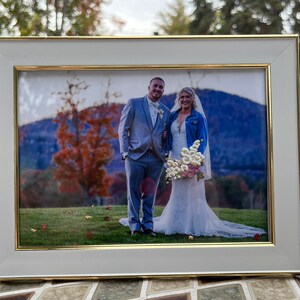 K&ouml;nnte beinhalten: Ein wei&szlig;er Bilderrahmen mit goldener Einfassung, der ein Hochzeitsfoto eines Paares zeigt, das vor einer Bergkette steht. Die Braut tr&auml;gt ein wei&szlig;es Kleid und eine Jeansjacke, der Br&auml;utigam tr&auml;gt einen grauen Anzug. Die Braut h&auml;lt einen Strau&szlig; wei&szlig;er Blumen.