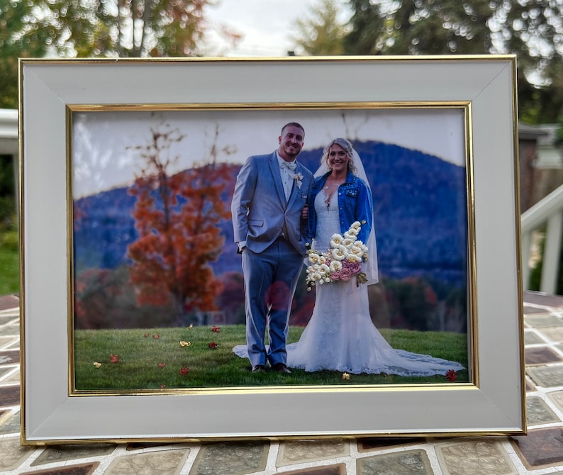 K&ouml;nnte beinhalten: Ein wei&szlig;er Bilderrahmen mit goldener Einfassung, der ein Hochzeitsfoto eines Paares zeigt, das vor einer Bergkette steht. Die Braut tr&auml;gt ein wei&szlig;es Kleid und eine Jeansjacke, der Br&auml;utigam tr&auml;gt einen grauen Anzug. Die Braut h&auml;lt einen Strau&szlig; wei&szlig;er Blumen.