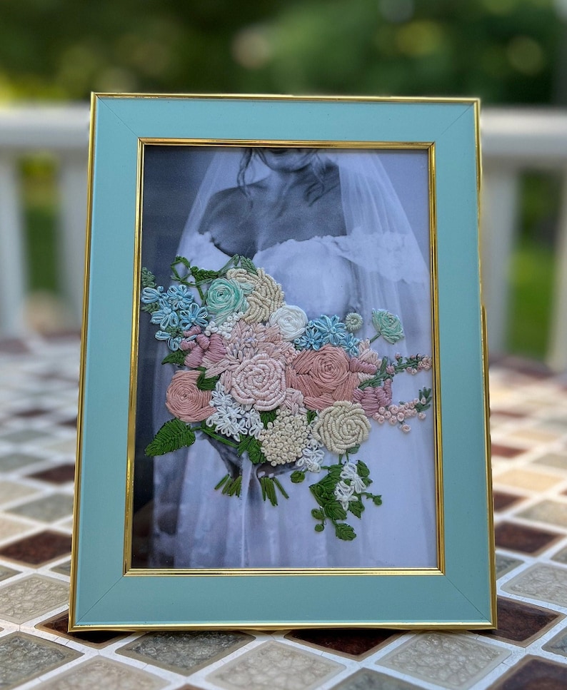 K&ouml;nnte beinhalten: Ein gerahmter gestickter Blumenstrau&szlig; in Rosa-, Wei&szlig;- und Blaut&ouml;nen mit gr&uuml;nen Bl&auml;ttern. Der Strau&szlig; befindet sich in einem hellblauen Rahmen mit goldener Verzierung.