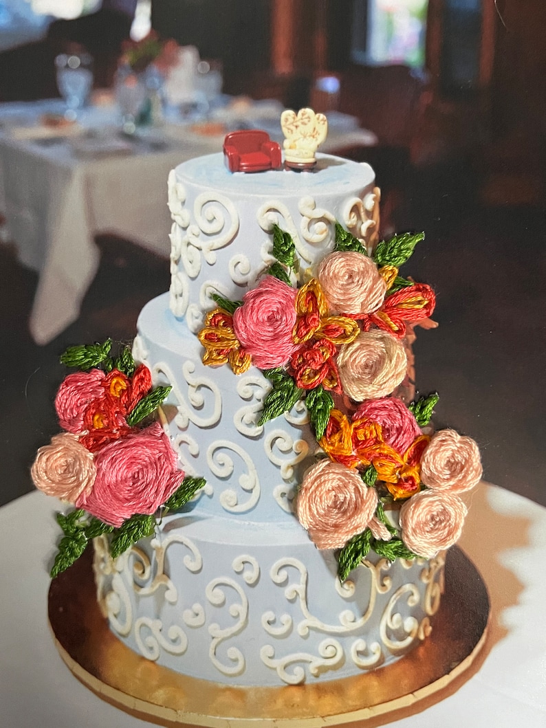 K&ouml;nnte beinhalten: Eine dreist&ouml;ckige Hochzeitstorte, verziert mit wei&szlig;em Frosting und filigranen Schn&ouml;rkeln. Die Torte ist mit einer Kaskade aus rosa, pfirsichfarbenen und orangefarbenen Stoffblumen geschm&uuml;ckt, die ein romantisches und elegantes Design schaffen. Ein kleines rotes Sofa und ein wei&szlig;er Sessel sind auf der Torte platziert und verleihen ihr einen verspielten Touch.