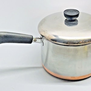 May include: Stainless steel saucepan with a copper-colored base and a lid. The pan has a black handle and a black knob on the lid. The pan is designed for cooking and is suitable for various kitchen tasks.